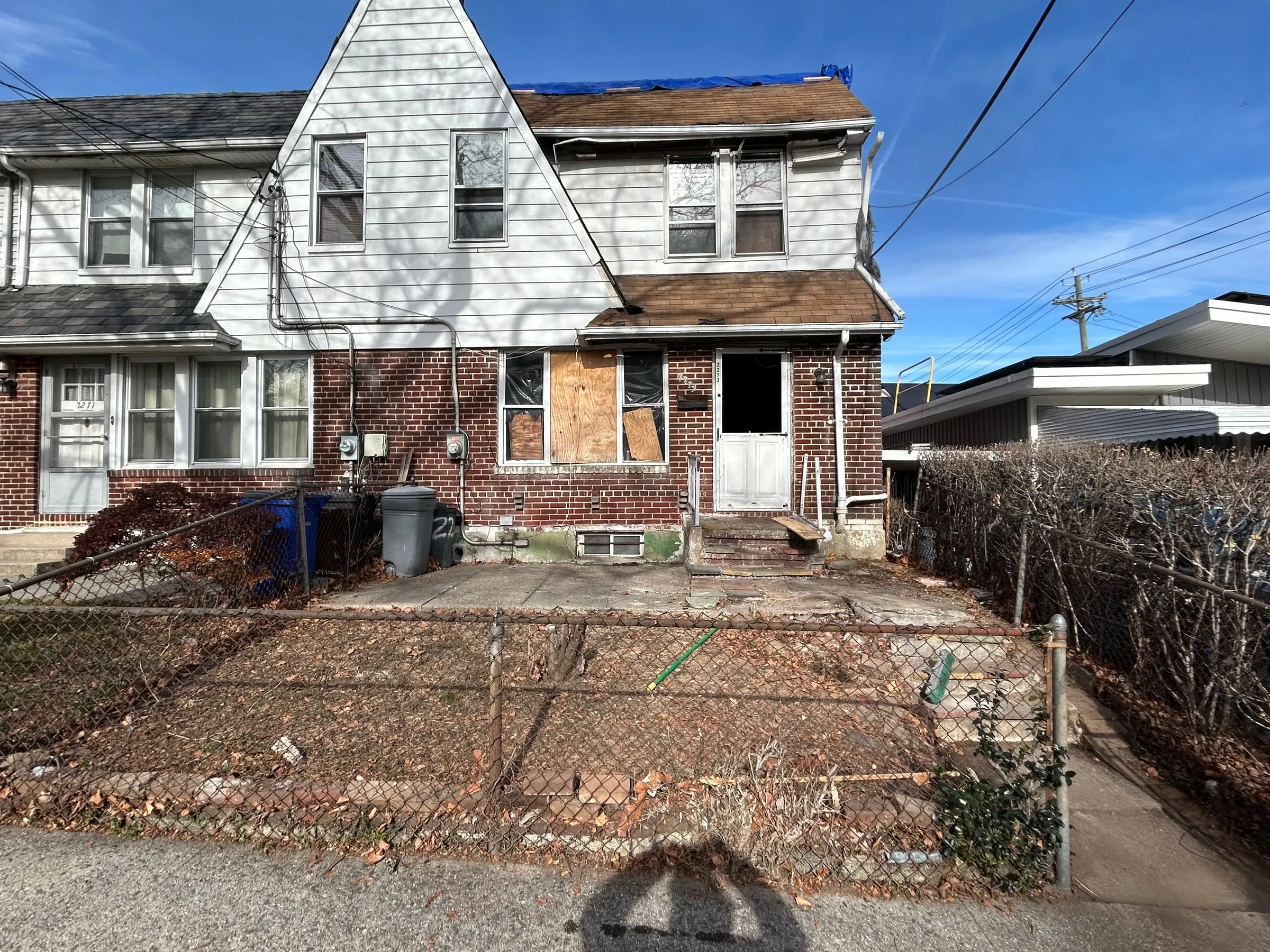 Boarded openings, a failed roof edge under tarpaulin, and a front yard behind chain link.